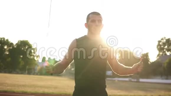 一名运动员穿着黑色运动服在体育场的跑道上跳过跳绳的手持镜头太阳视频的预览图