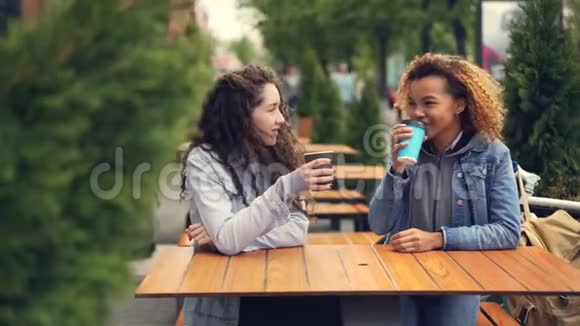 有魅力的白人和非裔美国女孩朋友正在城市的户外咖啡馆里喝咖啡聊天视频的预览图