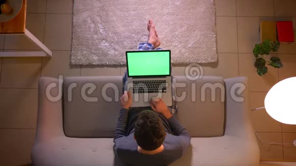 在舒适的室内沙发上坐着的时候用绿色屏幕在笔记本电脑上建立网络的特写镜头视频的预览图