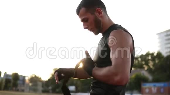一个年轻的留胡子的拳击手准备在户外训练一边裹着黑色绷带绷带一边在体育场上站着视频的预览图
