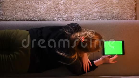 在舒适的公寓里躺在沙发上用绿色屏幕平板电脑拍摄漂亮女孩的特写镜头视频的预览图