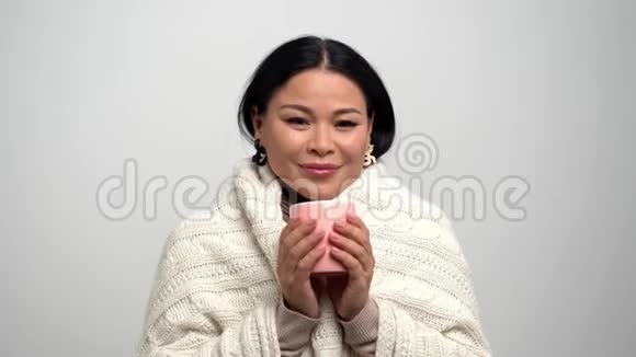 亚洲女人带着甜美的微笑裹着温暖的针织围巾喝着粉红色杯子里的茶视频的预览图