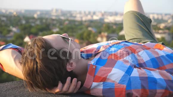 英俊的男人戴着太阳镜躺在屋顶上背景是城市景观年轻人在户外放松享受时光视频的预览图