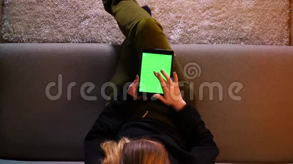 在舒适的室内沙发上坐着的时候用绿色屏幕在平板电脑上拍摄漂亮女孩的最新消息视频的预览图