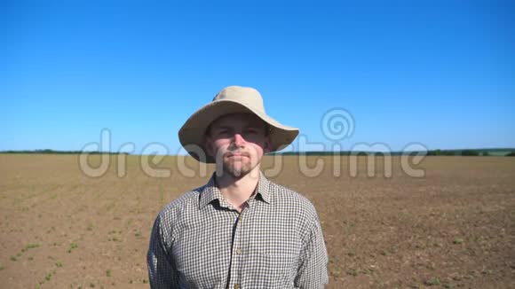 戴着帽子的快乐年轻农民的肖像看着镜头在田野和蓝天的背景下微笑视频的预览图