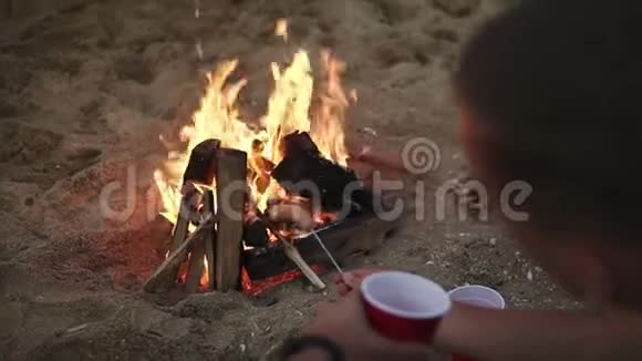 一对年轻的白种人夫妇正坐在海边的海滩上在棍子上烤大香肠视频的预览图