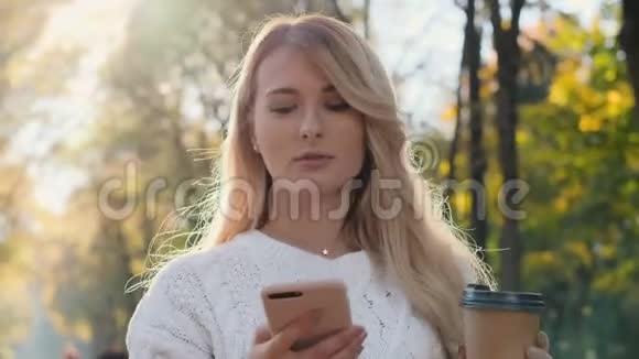 镜头跟随年轻迷人的时髦金发女孩用电话和喝咖啡去公园散步在柔软的公园视频的预览图