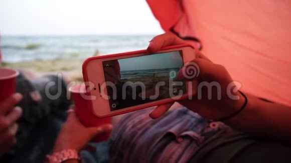 年轻夫妇在海边的红色帐篷里休息女人拿着智能手机拍两杯照片视频的预览图