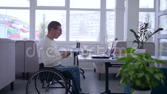 自由职业者男性残疾人戴着轮椅戴着眼镜工作在手机和电脑上坐在桌子旁视频的预览图
