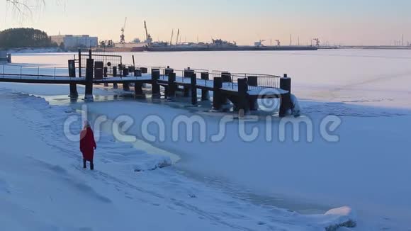穿着红色长夹克的年轻女子走在码头附近冰雪覆盖的河岸上视频的预览图