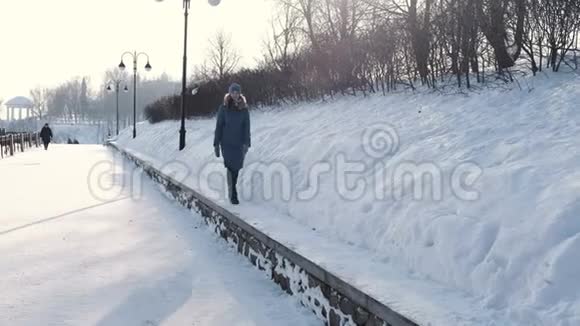 穿着蓝色羽绒服的年轻女子在冬季公园沿着边境散步看着天空前景视频的预览图