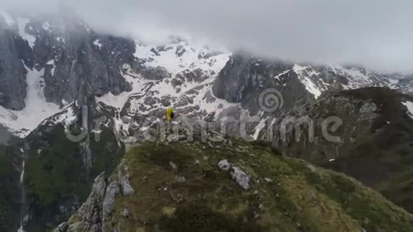 一个徒步旅行者站在山顶上看着白雪皑皑的山脉举起双手做胜利的手势视频的预览图