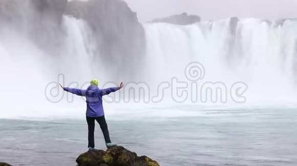 一个年轻的女人欣赏一个强大的狂暴瀑布沿着岩石边缘重重地落下一条晶莹清澈的冰川水流视频的预览图