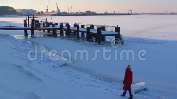 年轻女子穿着红色长袄走在码头附近冰雪覆盖的河岸上河港的背景视频的预览图