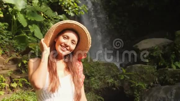 穿着白色连衣裙和草帽的年轻微笑混合种族旅游女孩与惊人的野生丛林瀑布的肖像生活方式视频的预览图