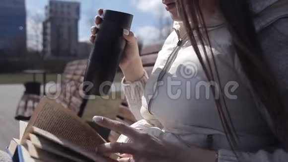 公园里的一个年轻女孩坐在长凳上看书喝着杯子里的咖啡近点视频的预览图