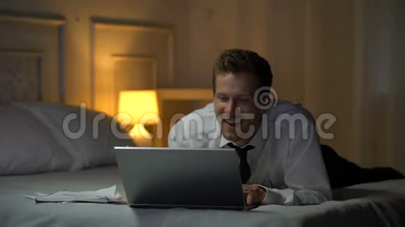 在床上微笑着穿着白色衬衫的男人在笔记本电脑上微笑着聊天办公室事务视频的预览图