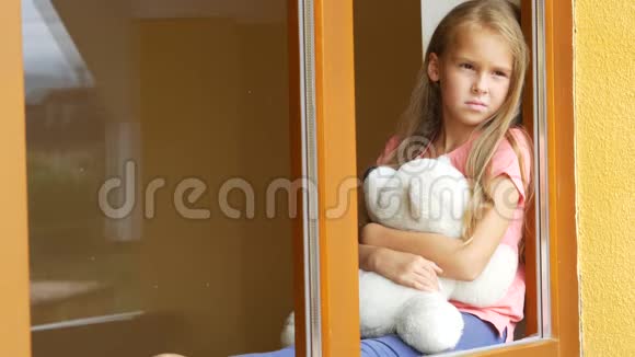 漂亮的小女孩带着柔软的玩具透过窗户看着视频的预览图