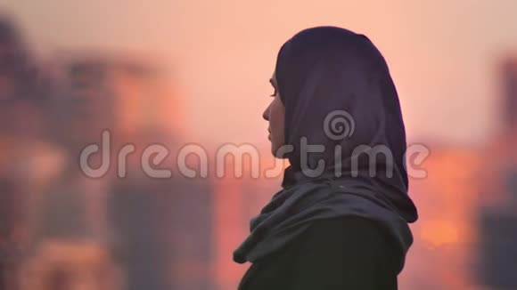 年轻迷人的穆斯林女性的特写肖像期待和转向相机与模糊的黄昏城市视频的预览图