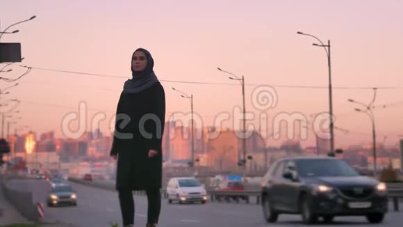 戴着头巾的年轻迷人的穆斯林女性站在过往的汽车旁与城市的镜头对视视频的预览图