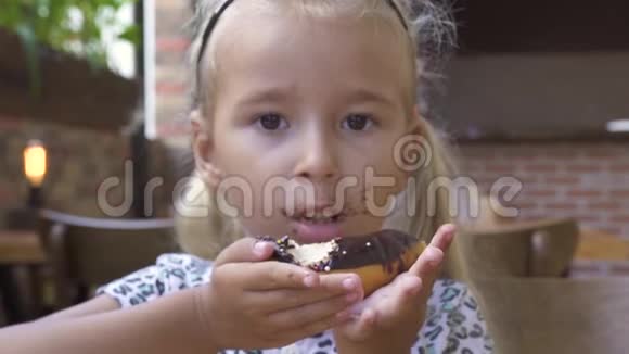 肖像小女孩在咖啡馆的桌子上吃甜巧克力甜甜圈可爱的金发女孩咬着巧克力甜甜圈视频的预览图