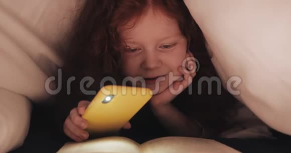 小红发女孩饶有兴趣地躺在他家的床上看书女孩用智能手机的手电筒视频的预览图