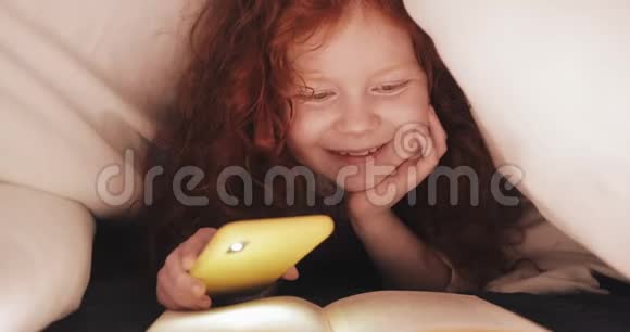 小红发女孩饶有兴趣地躺在他家的床上看书女孩用智能手机的手电筒视频的预览图
