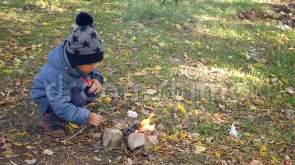 一个快乐的孩子正坐在森林里的炉火旁在一个凉爽的秋日和燃烧的棉花糖旅行和烹饪视频的预览图