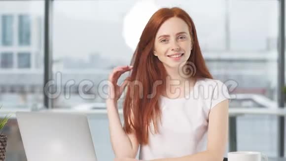 快乐的红发女孩坐在桌边拿着笔记本看着摄像机微笑的照片视频的预览图