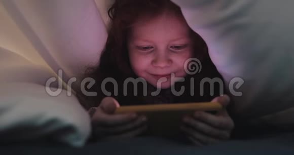红头发的小女孩晚上在黄色智能手机上看视频她躲在毯子下面视频的预览图