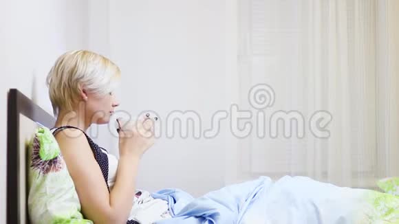 漂亮的金发女郎在床上喝咖啡用智能手机视频的预览图