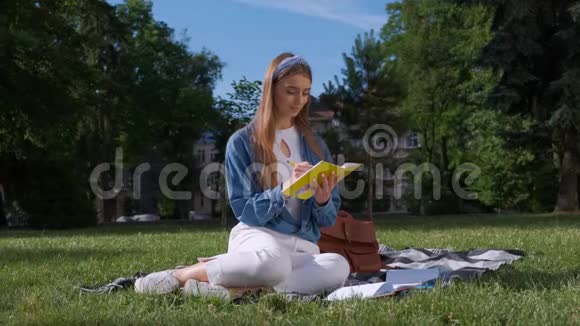 开朗的学生女孩学习她的笔记年轻女子坐在公园的草地上拿着一本打开的笔记本视频的预览图