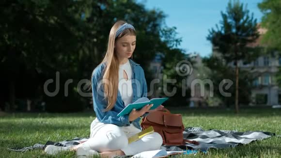 开朗的学生女孩学习她的笔记年轻女子坐在公园的草地上拿着一本打开的笔记本视频的预览图