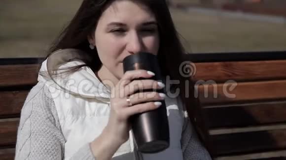 公园里的女孩坐在长凳上喝茶喝热杯子里的咖啡闭合躲避太阳视频的预览图