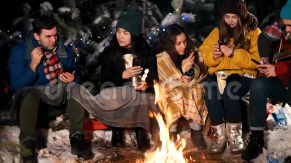 一群年轻人在冬林中坐在火边人们坐在手机上拍照视频的预览图
