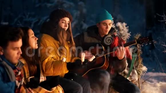 一群朋友坐在冬天的森林里在炉火旁吃棉花糖一个拿着吉他的年轻人视频的预览图