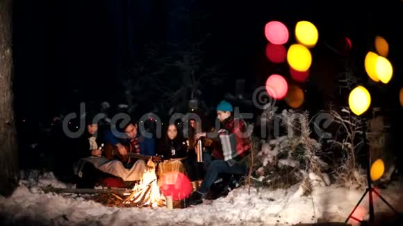 一群朋友坐在火堆旁的冬林里吃着棉花糖聊天拿吉他的年轻人视频的预览图