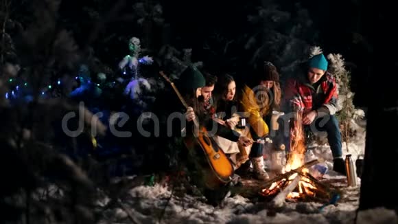 一群朋友坐在冬天的森林里在火边煎棉花糖一个拿着吉他的年轻人视频的预览图