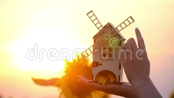 女孩手拿着玩具磨坊在夕阳和向日葵的背景下磨坊慢慢旋转的概念视频的预览图