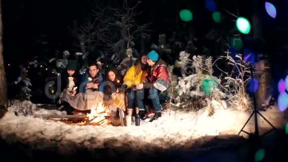冬天森林里坐在火堆旁的年轻人可爱的情侣之吻快乐的朋友们视频的预览图