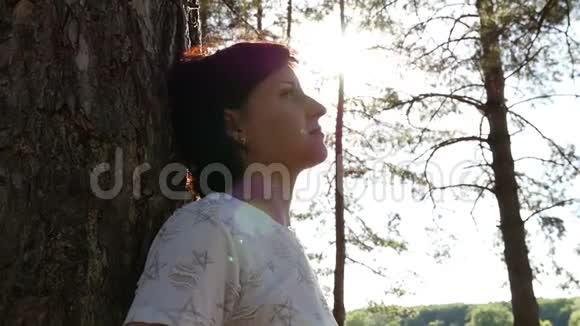 一位年轻的旅游妇女靠在森林里的一棵树上镜头围绕着森林里的女孩旋转休息视频的预览图