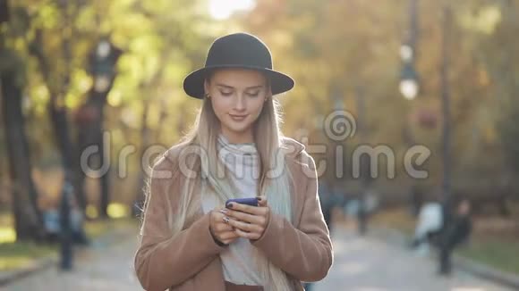 秋天城市公园散步时年轻漂亮的女士穿着时尚的外套戴着黑色的帽子使用智能手机视频的预览图