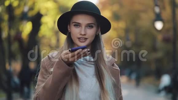 年轻漂亮的女士穿着时髦的外套戴着黑色帽子在秋天的公园里用智能手机发送语音信息视频的预览图
