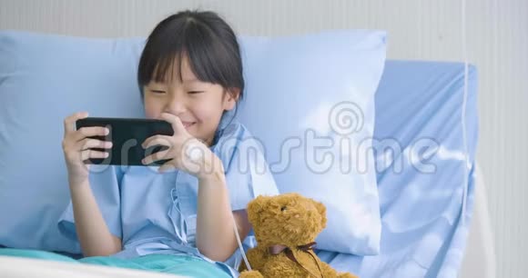 可爱的小女孩躺在医院的床上看有趣的卡通片智能手机上的电影疾病和治疗视频的预览图