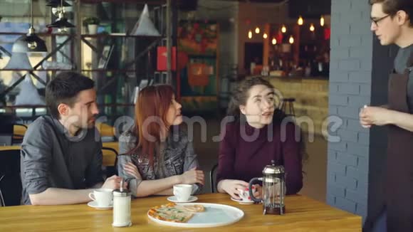 友好的男服务员正从一群快乐的朋友那里点餐他们坐在咖啡馆的桌子上聊天吃东西视频的预览图