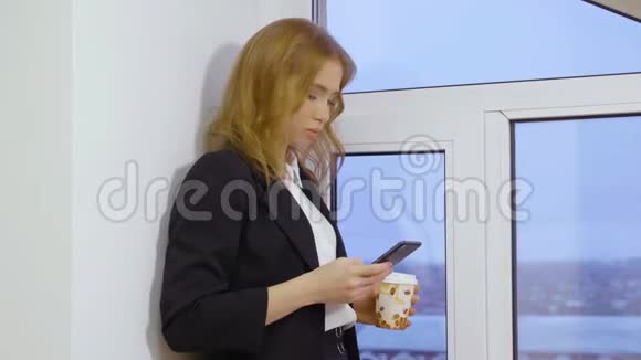 公司女经理在智能手机上发短信并在窗户附近端着一次性咖啡视频的预览图