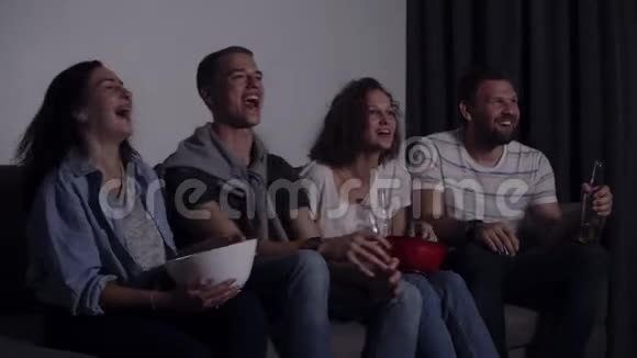四个朋友坐在沙发上在电视上看有趣的电影高加索年轻人坐在沙发上喝着饮料吃着零食电影视频的预览图