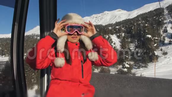 一个女孩坐在一个封闭的滑雪电梯上升到山顶女孩摘下滑雪护目镜享受美丽的冬天视频的预览图