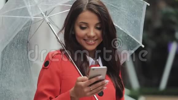 美丽的年轻商务女性在雨天在街上使用智能手机微笑着拿着雨伞拨号视频的预览图