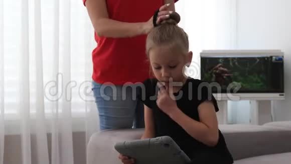 妈妈梳着可爱的女儿的头发她看着平板电脑妈妈和女儿在一起视频的预览图